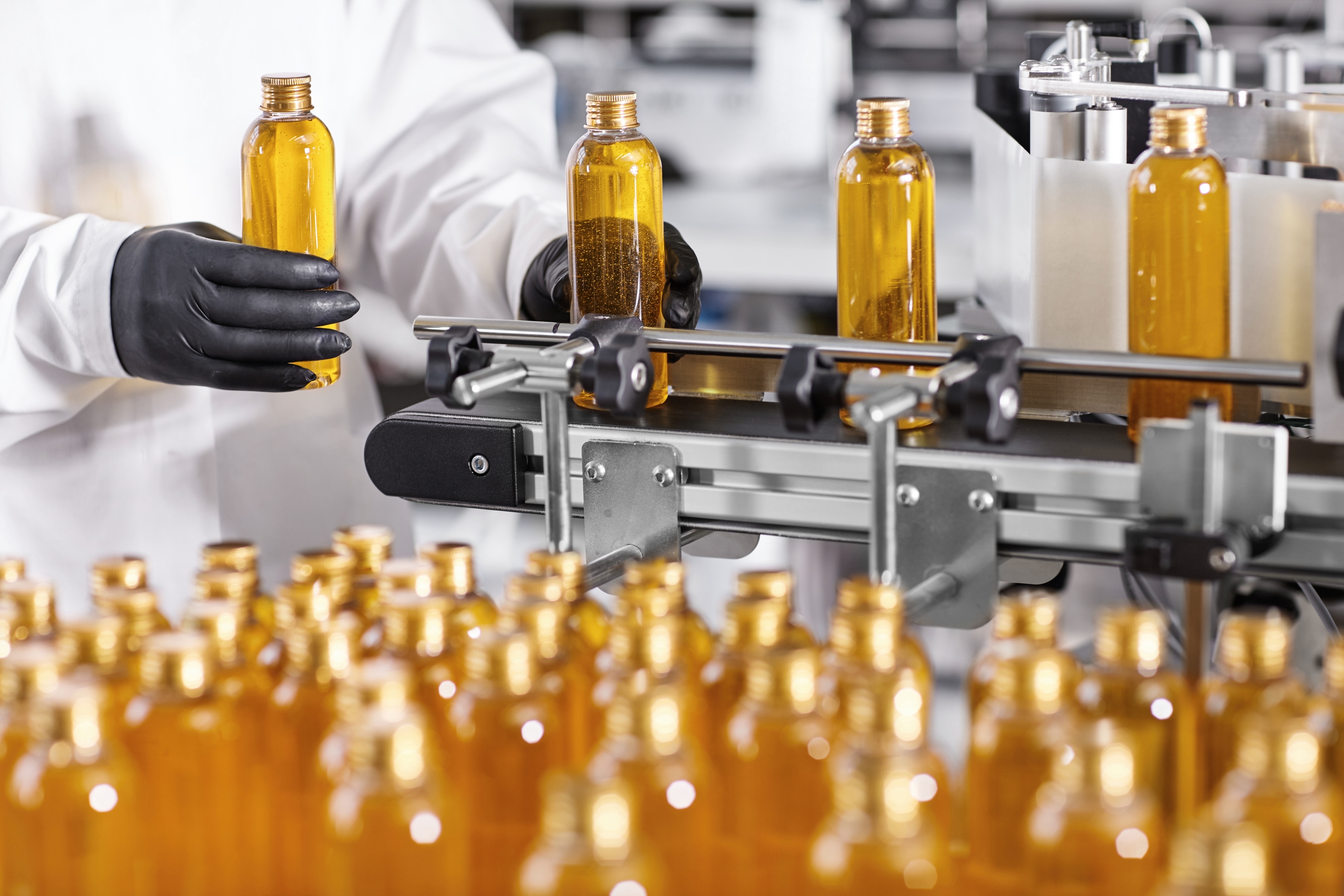 Plastic bottles with yellow liquid substance of shampoo, shower gel or soap standing in rows on assembly line at factory. Researcher in black gloves working at production line in modern laboratory